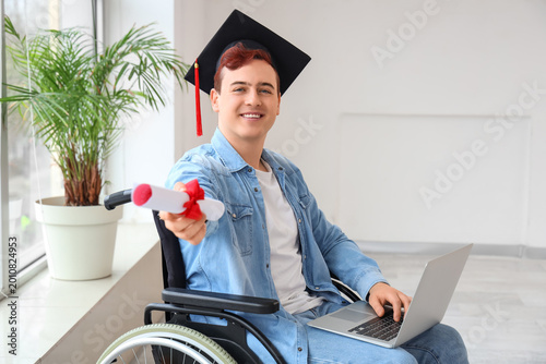 Male graduate in wheelchair with diploma and laptop at university hall
