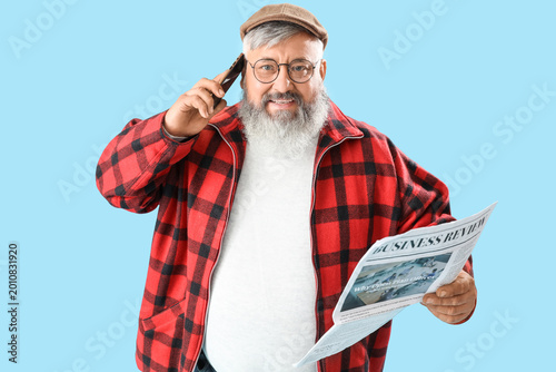 Mature man with newspaper talking by mobile phone on blue background