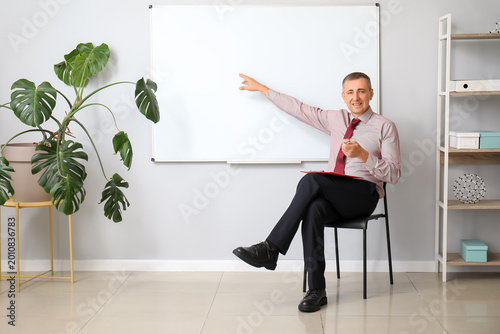 Male teacher with clipboard sitting on chair in classroom and pointing at fli...