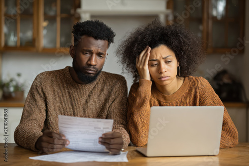 Young African American couple reviewing bills at kitchen table looking concerned while using laptop and paperwork in natural light.