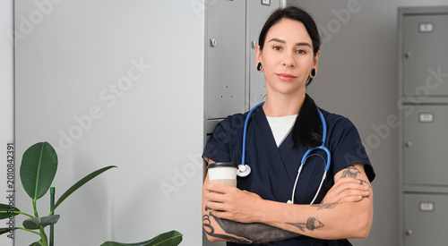Tattooed female doctor with coffee near locker in clinic