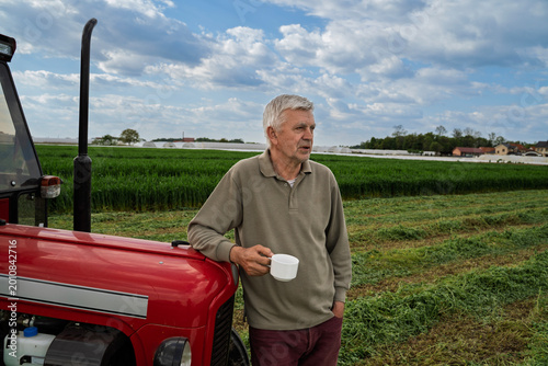 Fotografie Senior farmer on the filed leaned on a tractor with cup of coffee in his hands