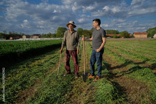 Two farmers senior and middle aged man standing on the agricultural field with tools