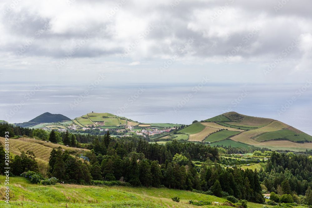 Obraz premium Atlantic Ocean view from San Miguel Island, Azores. Rolling hills and green fields with scattered houses. Moody sky with clouds over the island landscape.