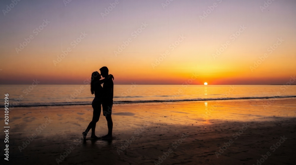 Obraz premium Silhouette of romantic couple embracing and kissing on beach at sunset. Romantic love, togetherness and summer vacation travel concept.