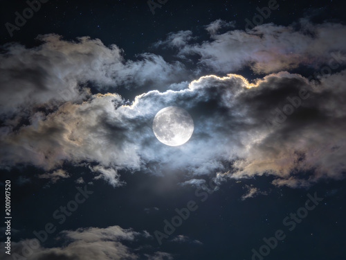 A breathtaking close up of a luminous full moon partially emerging from a dramatic swirl of thick textured clouds illuminated by soft golden light against a deep twilight sky showcasing