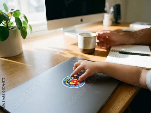 A child's small hand reaches for a colorful sticker on a closed laptop, resting on a sunlit wooden desk in a home office.