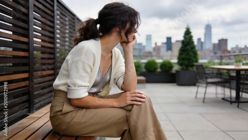 Pensive young woman sitting alone on a rooftop terrace overlooking the city skyline feeling sad lonely or deep in thought urban lifestyle concept