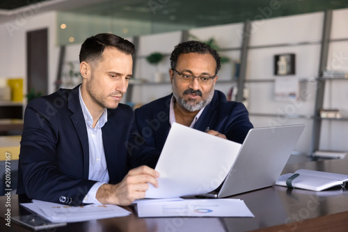 Fotografie Two focused business men colleagues reviewing financial statements and revenue report, analyzing printed charts, referencing data on laptop