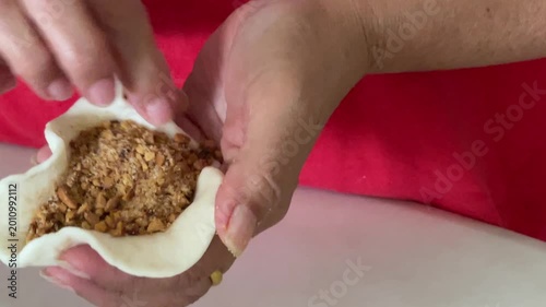 A close-up shot shows hands carefully stretching the edges of a dough wrapper to contain a very generous and overflowing amount of ground peanut filling for a handmade baozi.