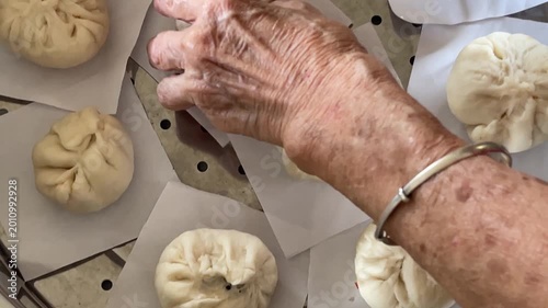 A close-up shot captures an elderly hand carefully placing a freshly pleat-wrapped raw baozi bun onto parchment paper inside a steamer tray, joining an arrangement of other handmade buns.