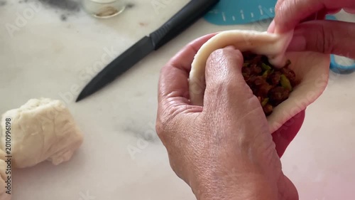 A close-up shows the hands of an elderly person pleating a handmade baozi, leaving a small opening at the top that reveals the savory minced meat and petai stuffing inside.