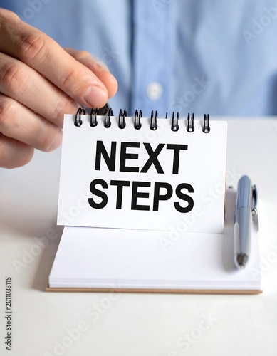 Close up of hand holding a pen near a notepad with next steps text