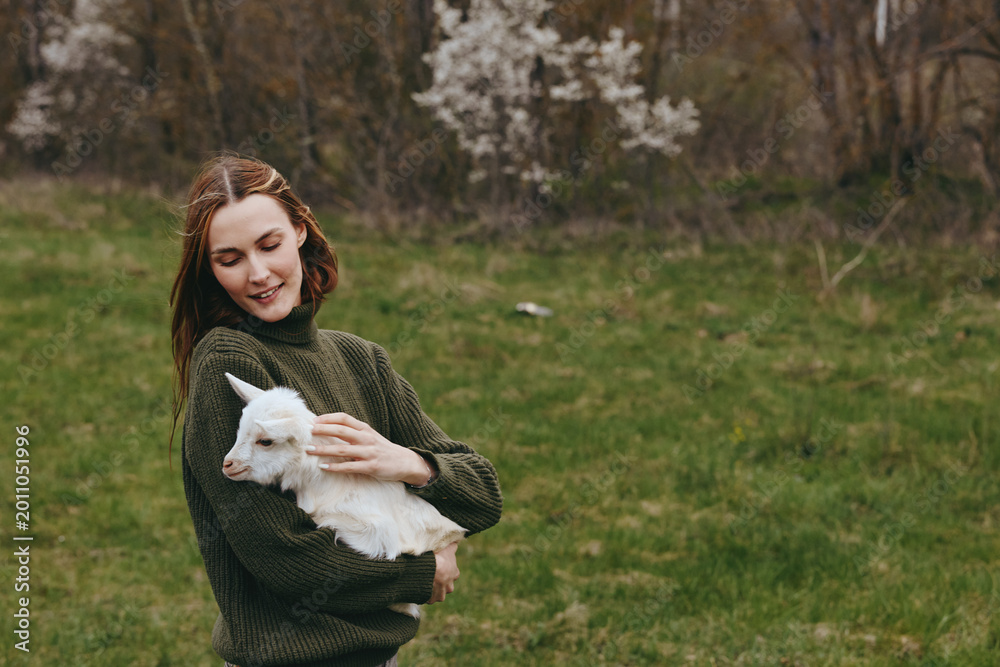 Fototapeta premium Gentle woman in a dark green sweater holds a white goat kid in a sunlit field, smiling softly, conveying calm rural lifestyle and close animal bond with open meadow behind, gentle breeze