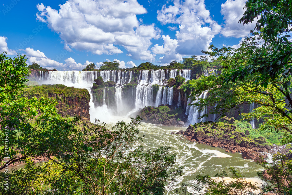 Naklejka premium Panoramic view of the majestic Iguazu Falls surrounded by lush green tropical rainforest under a blue cloudy sky, Misiones, Argentina