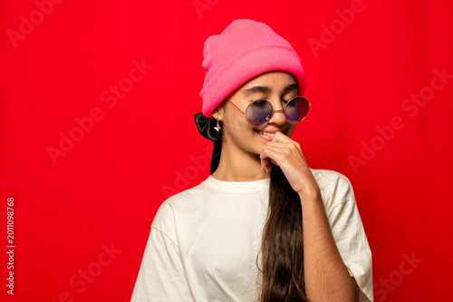 Young woman smiling wearing pink beanie and sunglasses in red background, studio shot