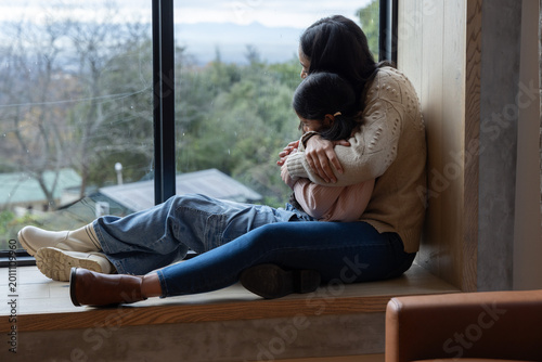Asian mother and child sitting on wooden bench by window hugging wearing sweater, jeans, copy space