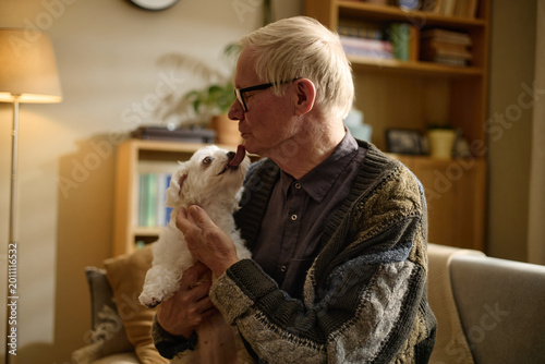 Fotografie Senior Caucasian man cuddling small dog at home, receiving affectionate lick and expressing companionship