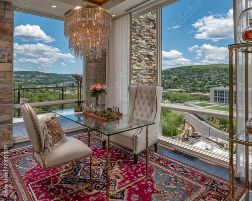 Glass desk stands elegantly in a modern sunroom decorated with a crystal chandelier and an ornate red Persian rug while providing a panoramic view of the lush green hillside and urban buildings.