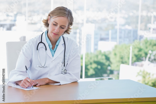 Schilderij op canvas Mature female doctor wearing white lab coat writing on paper at clinic desk with