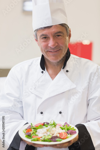 Mature chef wearing jacket and hat presenting plate with salad, halved tomatoes, protein in kitchen