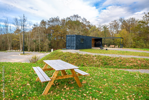 Wooden picnic table in front of modern lounge bar structure built from repurposed shipping containers, an outdoor terrace with seating arrangements and covered patio in peaceful natural green setting