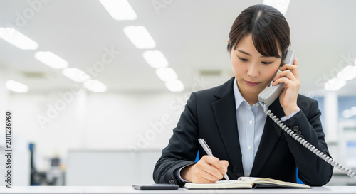 オフィスで電話をしながらメモを取る女性社員。アジア人、日本人。コピースペースあり
