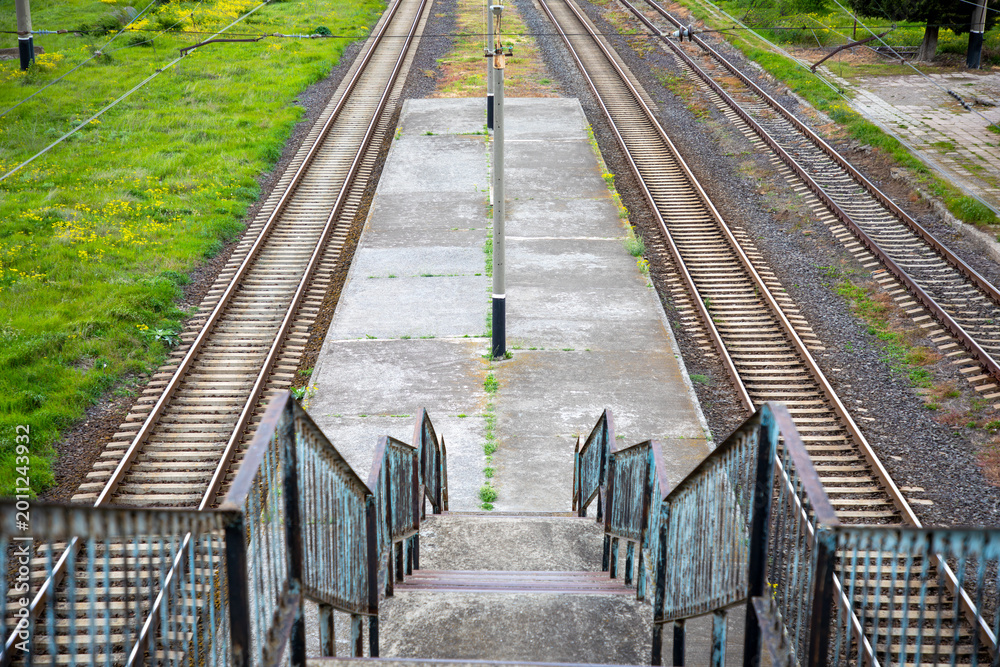 Obraz premium Concrete stairs leading down to an abandoned train station platform with parallel railway tracks stretching into the distance, conveying travel, journey, solitude, and destination