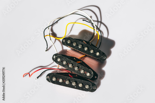 Electric guitar pickups with visible pole pieces are stacked together, each connected to colorful wires. Guitar transducers components rest on a white surface, casting subtle shadows