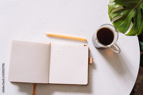 Open notebook with coffee and monstera leaf on table.
