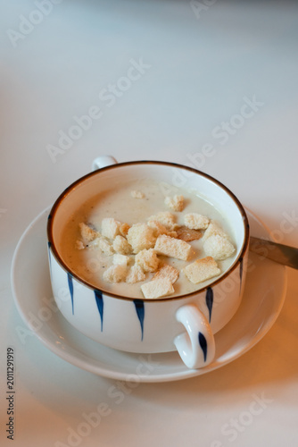 Close-up of a elegant serving of creamy soup in a stylish white ceramic cup with blue decorative patterns and a brown rim. The smooth, velvety soup is topped with golden-brown croutons and served on a