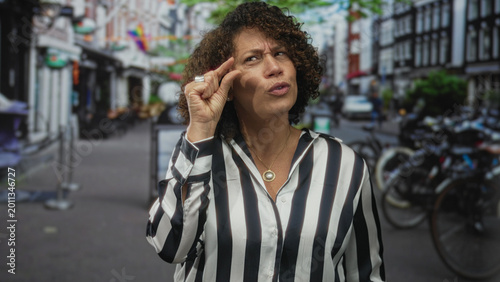 Fotografie Woman pinches fingers near her face on a busy urban street with bicycles and brick buildings in view; skepticism