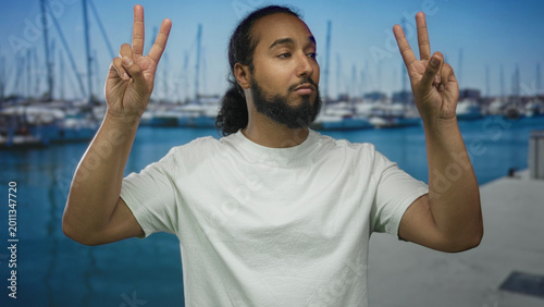 Φωτογραφία Man holding up two fingers in peace sign with both hands on a street side marina pier with visible boats and water; playful ease