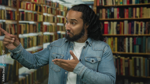 Bild auf Leinwand Man points finger to bookshelf in a library building while holding a book and gesturing with hand; skepticism curiosity study