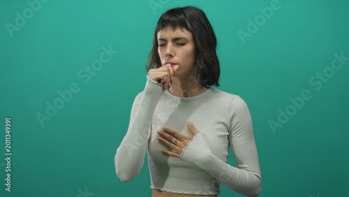 Fotografie Young hispanic woman coughs with hand on chest and eyes closed against green backdrop in studio; concern