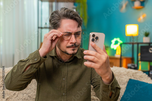 фотографія Middle-aged man at home in glasses squints reading tiny smartphone text worries about poor eyesight