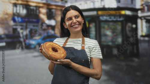 Photos Woman baker holds a round loaf with both hands, presenting the golden crusted bread while smiling on a busy city street near a kiosk and storefronts; pride