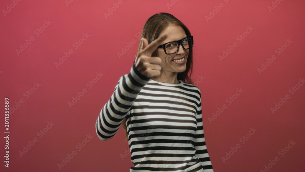 Obraz premium Woman wearing glasses and striped shirt smiles and raises fists in pink studio; joy playful celebration.