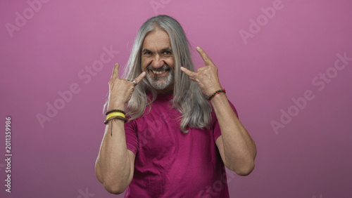 Fotografie Man middle age with hoary long hair grinning while showing rock horns hand gesture in pink studio with bracelets and rings visible; rebellion attitude energy