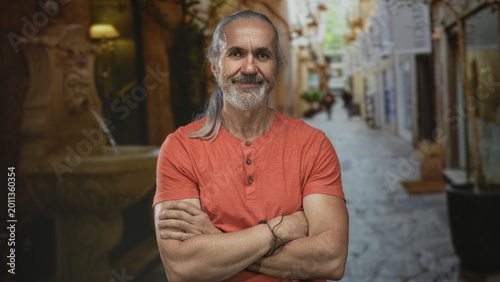 Fotografie Man with arms crossed showing forearms and slight smile in a cobblestone street near a fountain; confidence experience