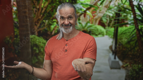 Foto Man with long gray hair and beard holds both palms up on a leafy park path in forest, smiling gently; serenity