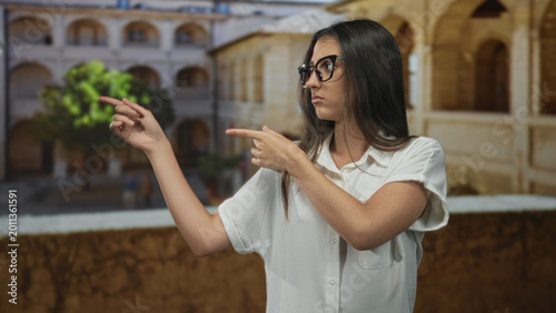 Foto Woman points fingers to the left on an old town street, wearing glasses and white shirt, bare forearms visible; skepticism doubt indecision