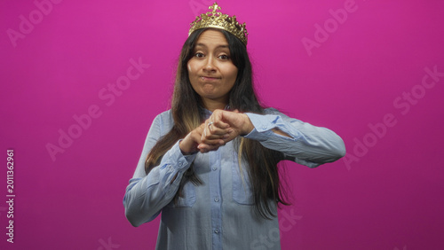 Foto Young latina woman wearing gold crown and blue shirt taps her wrist and gestures checking time in magenta studio; impatience