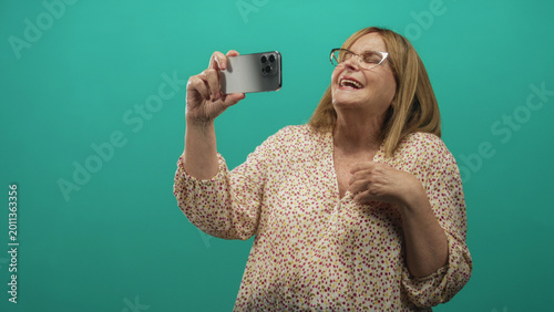 Obraz na płótnie Senior woman wearing glasses and floral blouse holds smartphone with raised arm and hand on chest in studio; joy connection
