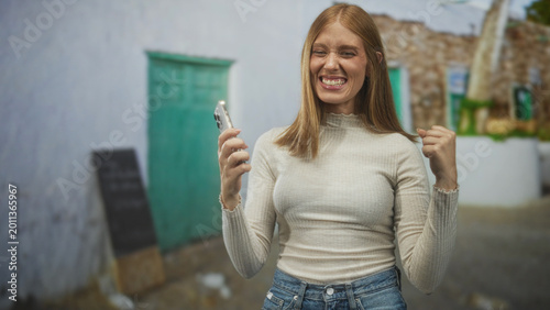 Fotografía Woman holding smartphone, smiling and pumping fist while later speaking on phone on a street with green doors and stone walls of a village; celebration joy