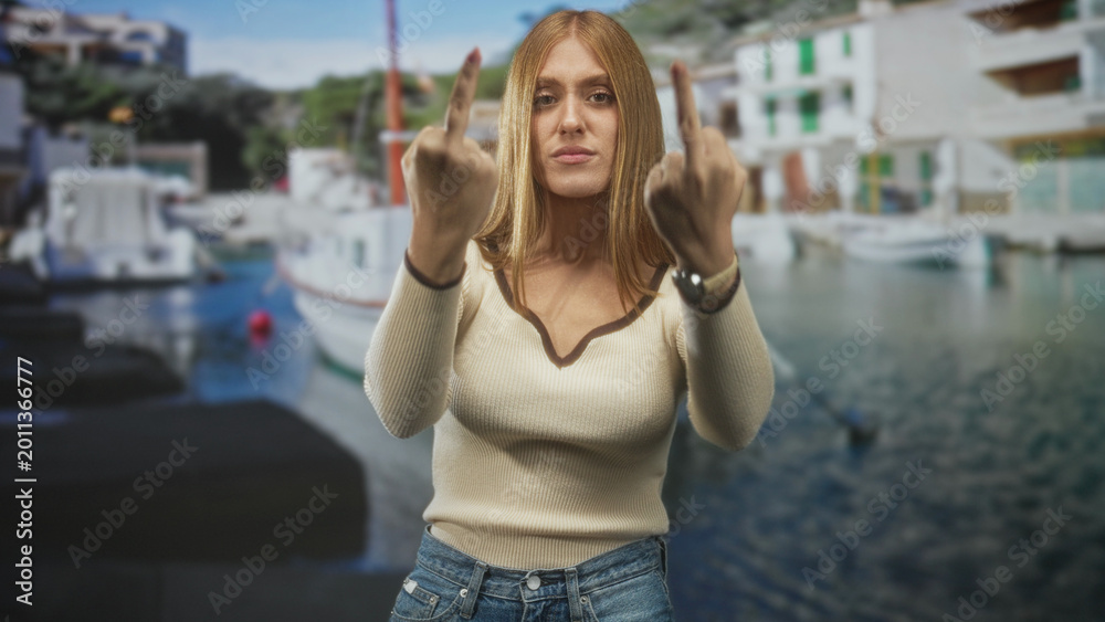 Fototapeta premium Woman holding up middle fingers at a harbor pier, redhead in cream sweater and denim jeans with wristwatch visible; defiance attitude.