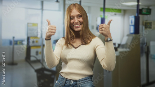 Fotografía Woman giving thumbs up and smiling at airport checkin kiosk among monitors and queue stanchions, wearing sweater and jeans; joy confidence