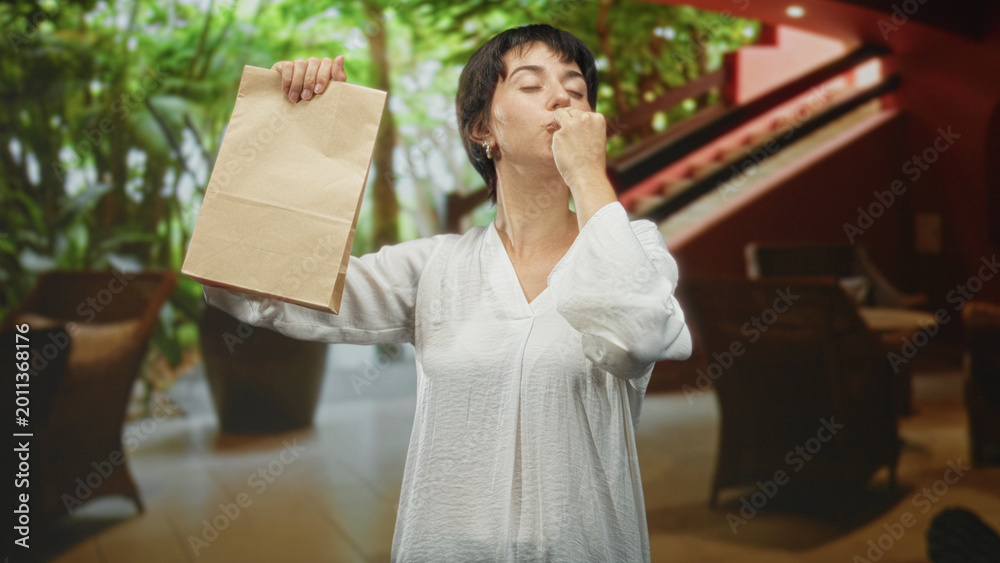 Fototapeta premium Woman sniffs brown paper bag while raising one hand and smiling in a building lobby with plants and chairs; delight discovery.