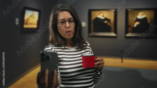 Bild auf Leinwand Woman brunette middle aged holding red mug and smartphone makes puckered lips selfie in museum building among framed paintings; playful reflection