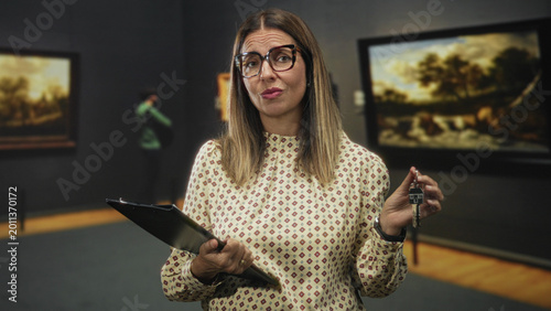 Obraz na plátně Middle age woman holds keys and clipboard and makes a skeptical face while standing in a museum building; skepticism